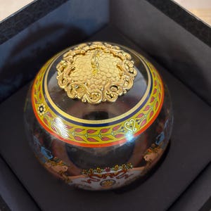 Puede incluir: Una caja decorativa redonda con base negra y tapa dorada ornamentada. La caja presenta una banda colorida con motivos florales y geom&eacute;tricos. La caja se presenta dentro de una caja negra.