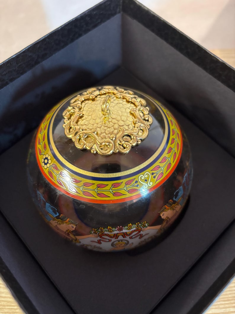 Puede incluir: Una caja decorativa redonda con base negra y tapa dorada ornamentada. La caja presenta una banda colorida con motivos florales y geom&eacute;tricos. La caja se presenta dentro de una caja negra.