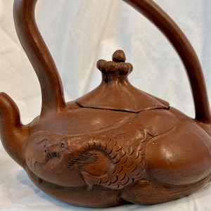 May include: A brown clay teapot with a dragon design. The teapot has a curved handle and a lid with a knob on top.
