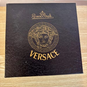Puede incluir: Una caja negra con el logotipo de Versace en dorado. El logotipo presenta una cabeza de Medusa dentro de un marco circular. La palabra "VERSACE" est&aacute; impresa en dorado debajo. La caja tambi&eacute;n tiene el logotipo "Rosenthal".
