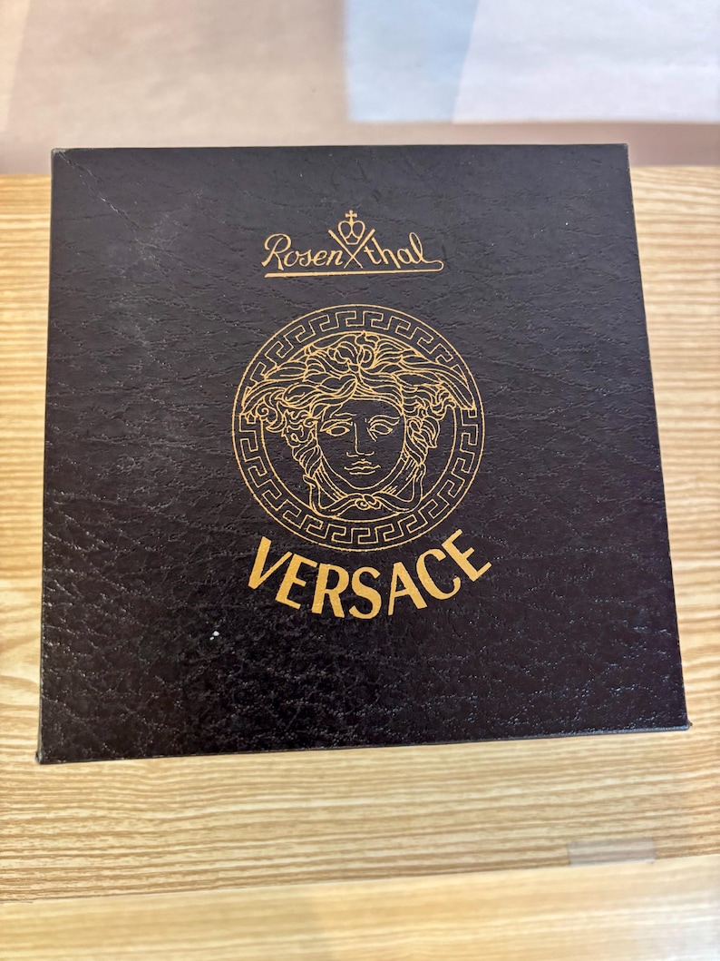 Puede incluir: Una caja negra con el logotipo de Versace en dorado. El logotipo presenta una cabeza de Medusa dentro de un marco circular. La palabra "VERSACE" est&aacute; impresa en dorado debajo. La caja tambi&eacute;n tiene el logotipo "Rosenthal".