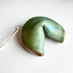 May include: A ceramic fortune cookie ornament with a glossy, mottled green glaze on the exterior and a textured, unglazed brown clay finish on the interior. A thin metal wire loop is attached to one side, connected to a light tan twine string.