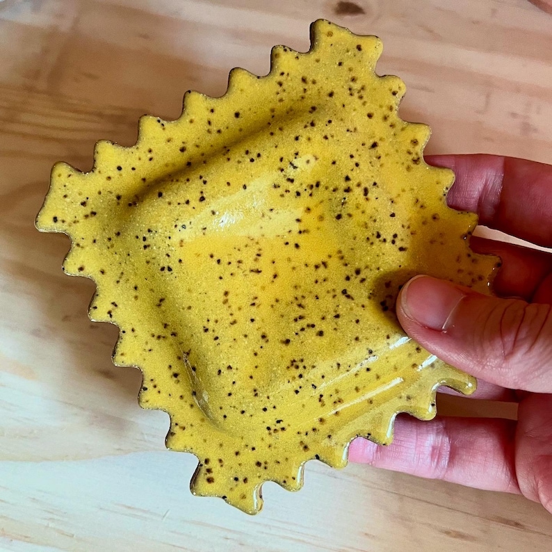 May include: A handmade, yellow ceramic ravioli-shaped dish with a speckled pattern. The dish has a scalloped edge and a recessed center. The ceramic dish is held in a person's hand.