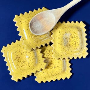 May include: A wooden spoon rests on three yellow ceramic ravioli-shaped dishes with speckled detailing. The dishes have a wavy edge and are set against a deep blue background. Kitchenware.
