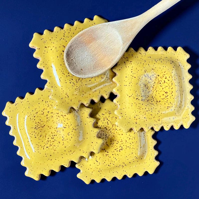May include: A wooden spoon rests on three yellow ceramic ravioli-shaped dishes with speckled detailing. The dishes have a wavy edge and are set against a deep blue background. Kitchenware.