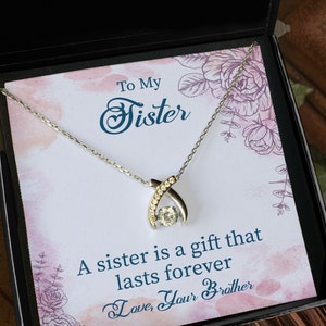 Pode incluir: Um colar de prata com um pingente de cristal transparente em uma caixa de presente preta. A caixa tem um design floral e o texto "To My Sister A sister is a gift that lasts forever Love, Your Brother".
