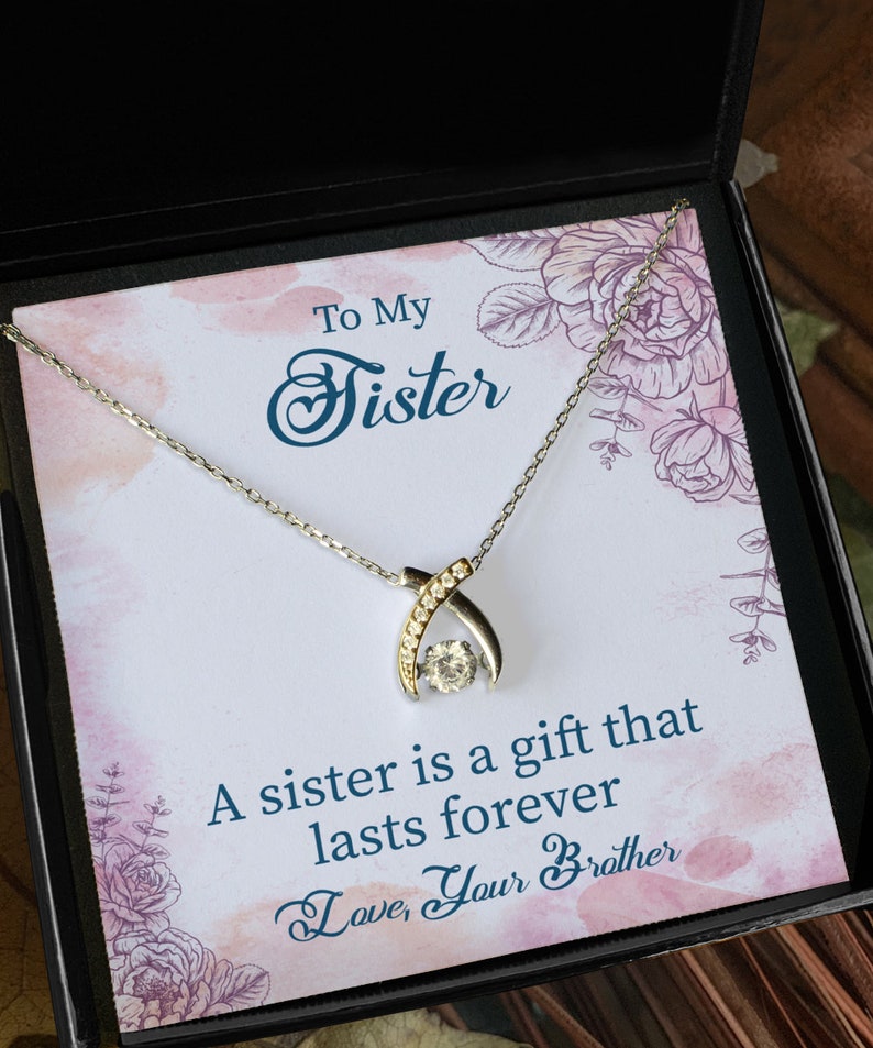Pode incluir: Um colar de prata com um pingente de cristal transparente em uma caixa de presente preta. A caixa tem um design floral e o texto "To My Sister A sister is a gift that lasts forever Love, Your Brother".