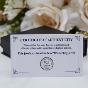 K&ouml;nnte beinhalten: Eine wei&szlig;e Karte mit schwarzem Rand und dem aufgedruckten Text "CERTIFICATE OF AUTHENTICITY". Die Karte bescheinigt, dass der Schmuck authentisch ist und aus 925er Sterlingsilber handgefertigt ist. Ein rundes Siegel mit dem Text "SEAL OF AUTHENTICITY 925 Sterling Silver" ist auf die Karte gedruckt.