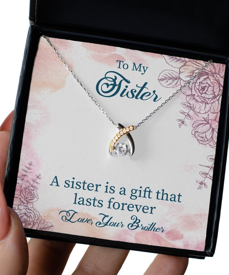 Pode incluir: Um colar de prata com um pingente de cristal transparente em uma caixa com um design floral rosa e branco. A caixa tem uma mensagem que diz "To My Sister A sister is a gift that lasts forever Love, Your Brother".