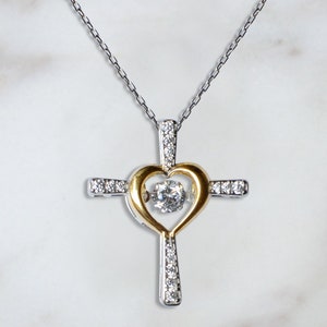 K&ouml;nnte beinhalten: Eine silberne Kettenkette mit einem silbernen und goldenen Kreuzanh&auml;nger. Das Kreuz hat eine herzf&ouml;rmige &Ouml;ffnung mit einem klaren Kristall in der Mitte.