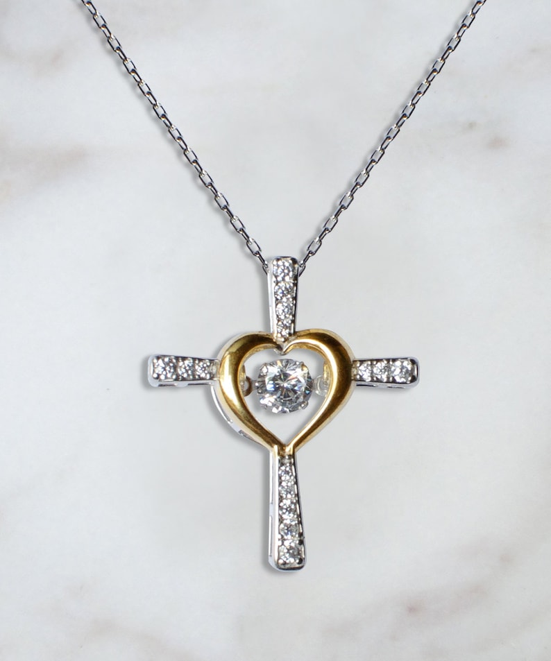 K&ouml;nnte beinhalten: Eine silberne Kettenkette mit einem silbernen und goldenen Kreuzanh&auml;nger. Das Kreuz hat eine herzf&ouml;rmige &Ouml;ffnung mit einem klaren Kristall in der Mitte.
