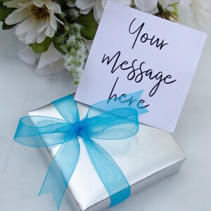 Pode incluir: Uma caixa de presente prateada com uma fita e um la&ccedil;o azuis. Um cart&atilde;o branco com o texto "Your message here" est&aacute; em cima da caixa.