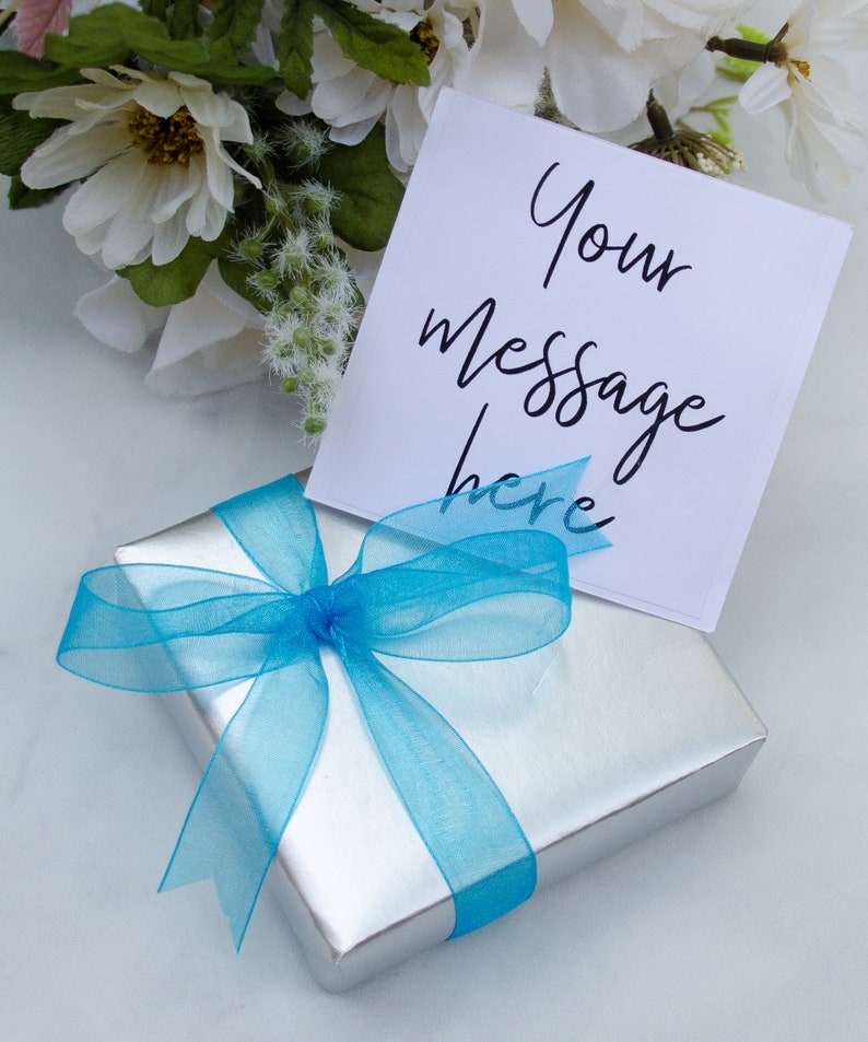 Pode incluir: Uma caixa de presente prateada com uma fita e um la&ccedil;o azuis. Um cart&atilde;o branco com o texto "Your message here" est&aacute; em cima da caixa.