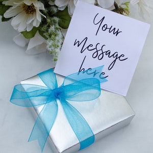 K&ouml;nnte beinhalten: Eine silberne Geschenkbox mit einem blauen Band und Schleife. Eine wei&szlig;e Karte mit dem Text "Your message here" liegt auf der Box.
