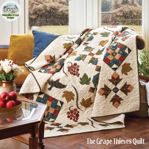 May include: A cream-coloured quilt with a patchwork design featuring a variety of fabrics in shades of red, green, blue, and brown. The quilt is draped over a sofa and features a design of grapes, leaves, and birds. The quilt is titled "The Grape Thieves Quilt".
