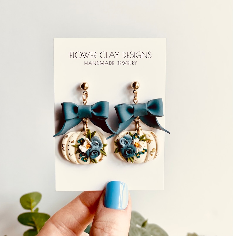 May include: Handmade earrings with a white pumpkin design, blue floral accents, and a teal bow. The earrings hang from gold-coloured posts. The card behind the earrings reads "FLOWER CLAY DESIGNS HANDMADE JEWELRY."