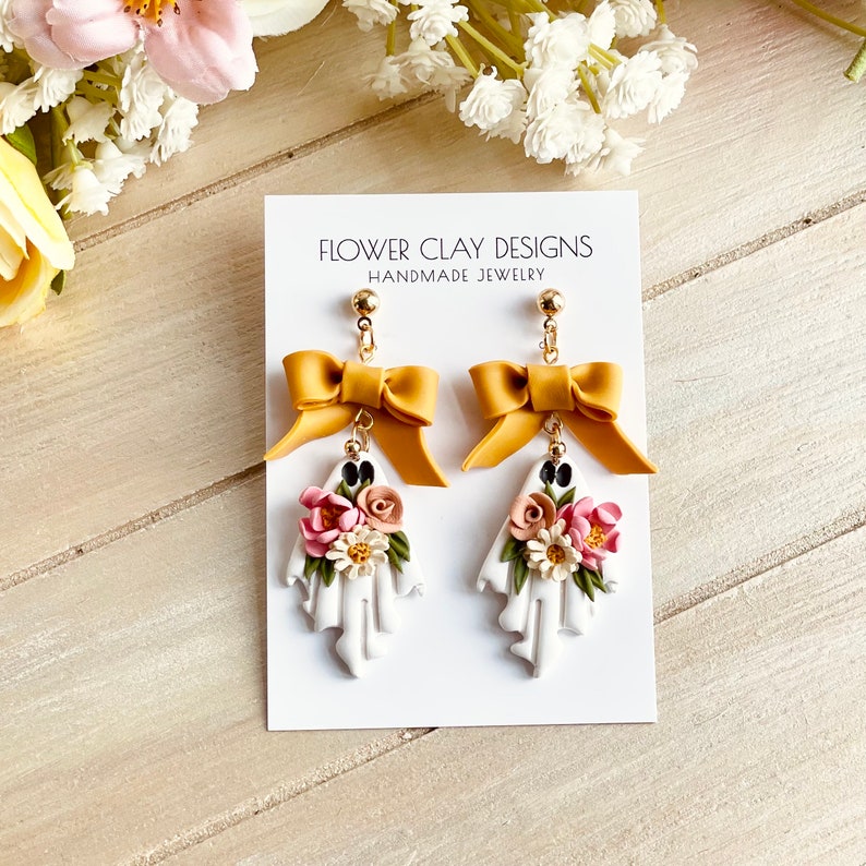 May include: A pair of handmade earrings featuring a white ghost design with floral accents and a mustard yellow bow. The earrings are attached to a gold-colored stud. The card behind the earrings reads "FLOWER CLAY DESIGNS HANDMADE JEWELRY."