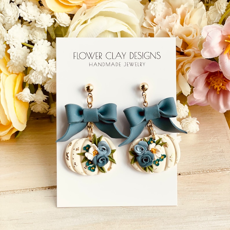 May include: Handmade earrings with a bow and pumpkin design. The earrings have a teal bow, a gold chain, and a white pumpkin with blue and white flowers. The card reads "FLOWER CLAY DESIGNS HANDMADE JEWELRY."
