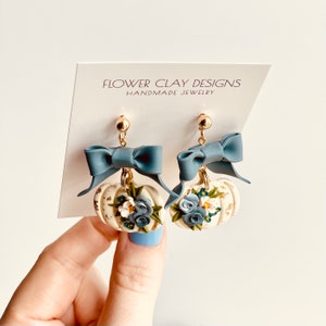 May include: Handmade earrings with a pumpkin design. The cream-coloured pumpkins have gold accents, blue and white flowers, and a blue bow. The earrings hang from gold-coloured posts. The card behind the earrings reads "FLOWER CLAY DESIGNS HANDMADE JEWELRY."