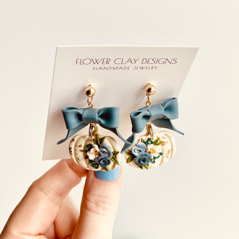 May include: Handmade earrings with a pumpkin design. The cream-coloured pumpkins have gold accents, blue and white flowers, and a blue bow. The earrings hang from gold-coloured posts. The card behind the earrings reads "FLOWER CLAY DESIGNS HANDMADE JEWELRY."