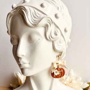 K&ouml;nnte beinhalten: Herbstliche Ohrringe mit einem K&uuml;rbis-Design. Die Ohrringe sind warmbraun mit wei&szlig;en floralen Akzenten und goldenen Details. Jeder Ohrring hat eine cremefarbene Schleife und einen goldfarbenen Stecker.