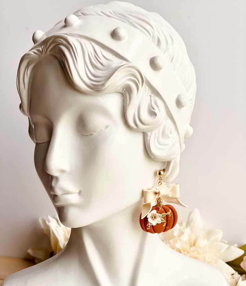K&ouml;nnte beinhalten: Herbstliche Ohrringe mit einem K&uuml;rbis-Design. Die Ohrringe sind warmbraun mit wei&szlig;en floralen Akzenten und goldenen Details. Jeder Ohrring hat eine cremefarbene Schleife und einen goldfarbenen Stecker.