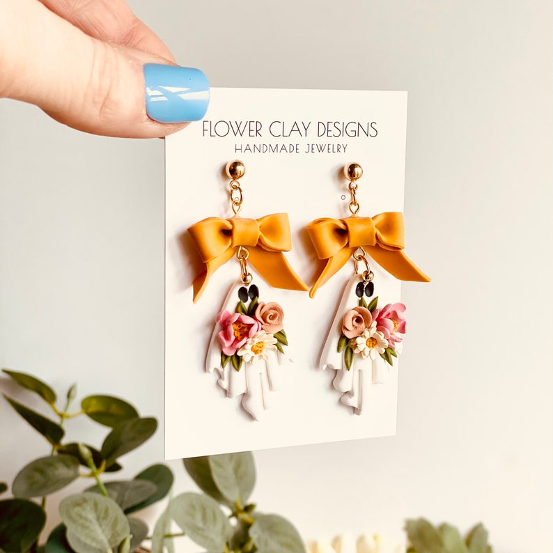 May include: A pair of handmade earrings featuring a ghost design with a yellow bow and floral accents. The earrings are attached to a card that reads "FLOWER CLAY DESIGNS HANDMADE JEWELRY". The earrings are a unique accessory.