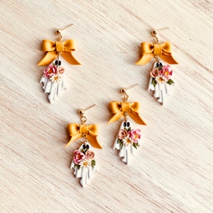 May include: Four pairs of handmade ghost earrings. Each earring features a white ghost shape with a floral arrangement and a gold bow. The earrings have gold-colored posts.