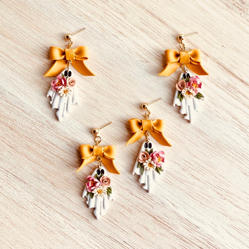 May include: Four pairs of handmade ghost earrings. Each earring features a white ghost shape with a floral arrangement and a gold bow. The earrings have gold-colored posts.