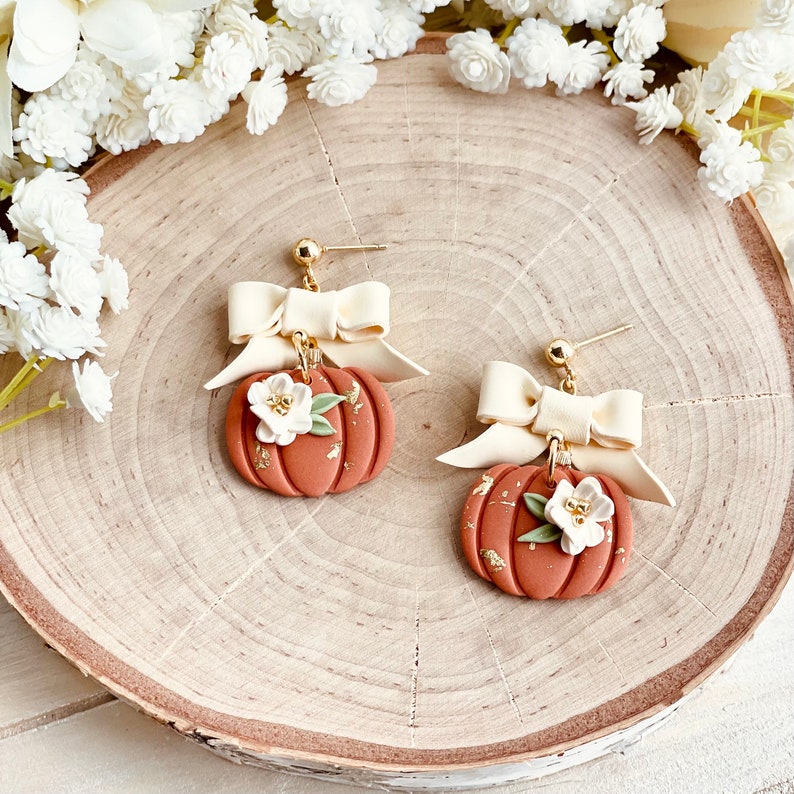 K&ouml;nnte beinhalten: Ohrringe in K&uuml;rbisform in Terrakotta mit cremefarbenen Schleifen und wei&szlig;en Blumen. Die Ohrringe haben goldene Ohrstecker und Goldflitter. Ein herbstliches, saisonales Design.