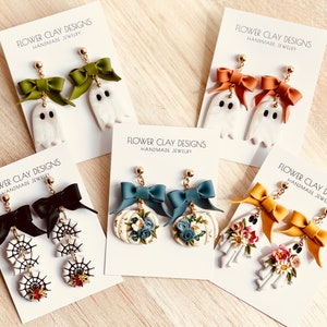 May include: Handmade Halloween earrings with ghost, spiderweb, pumpkin, and skeleton designs. The earrings have colourful bows and floral accents. The cards display the text "FLOWER CLAY DESIGNS HANDMADE JEWELRY".