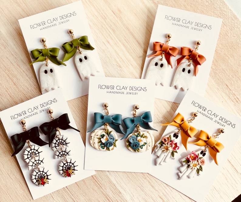 May include: Handmade Halloween earrings with ghost, spiderweb, pumpkin, and skeleton designs. The earrings have colourful bows and floral accents. The cards display the text "FLOWER CLAY DESIGNS HANDMADE JEWELRY".