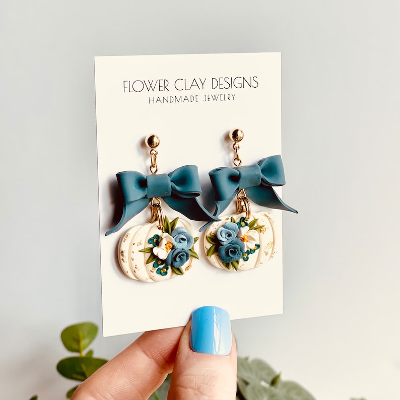 May include: Handmade earrings with white pumpkin charms decorated with blue and white flowers and gold details. Each earring has a teal bow and a gold stud. The card behind the earrings reads "FLOWER CLAY DESIGNS HANDMADE JEWELRY".