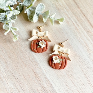 K&ouml;nnte beinhalten: Handgefertigte Ohrringe mit K&uuml;rbis-Design. Die Ohrringe sind in einem warmen Braunton gehalten und haben eine cremefarbene Schleife, eine wei&szlig;e Blume und goldfarbene Akzente. Die Ohrringe sind mit einem Stecker versehen.