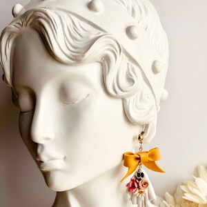 May include: Mustard yellow bow earrings with a small bouquet of pink, white, and black flowers. The earrings are hanging from a gold stud. The earrings are displayed on a white bust of a woman.