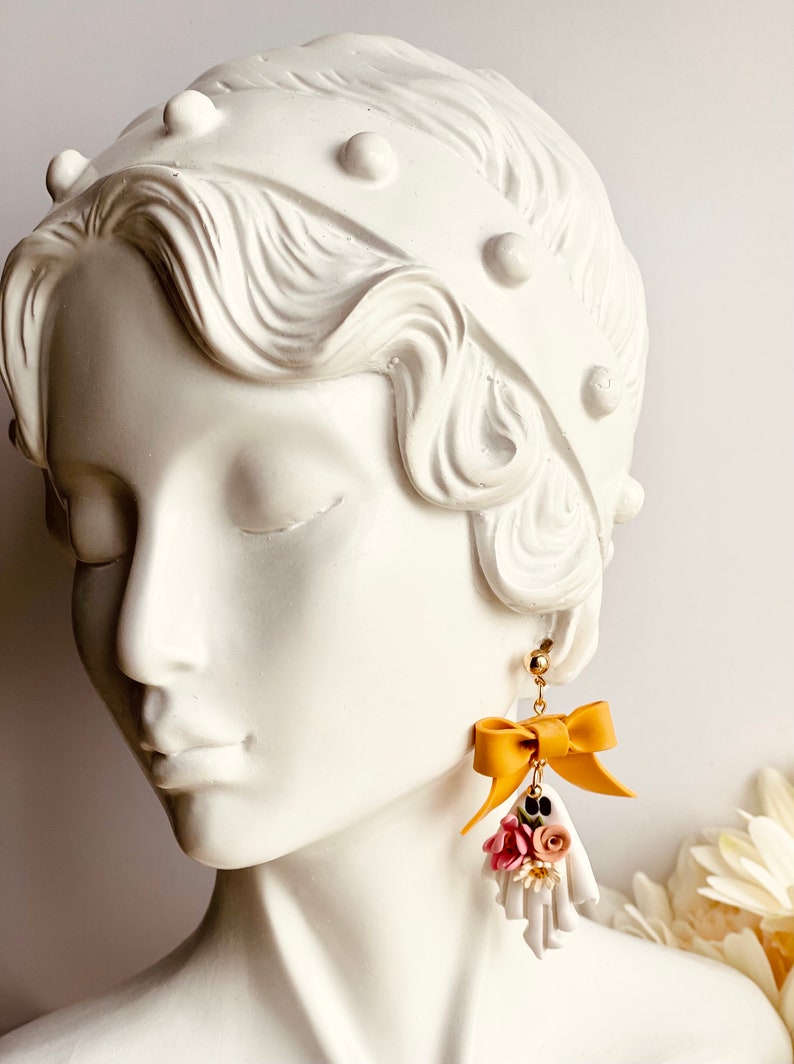 May include: Mustard yellow bow earrings with a small bouquet of pink, white, and black flowers. The earrings are hanging from a gold stud. The earrings are displayed on a white bust of a woman.