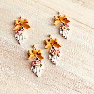 May include: Four pairs of white ghost-shaped earrings with gold-colored bows and floral accents. The earrings have gold-colored posts and are arranged on a light wood-grain surface. The flowers are pink, white, and brown.