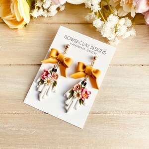 May include: A pair of handmade ghost earrings with a floral arrangement and a mustard yellow bow. The earrings are white with a floral arrangement of pink, white, and green flowers. The text on the card reads "FLOWER CLAY DESIGNS HANDMADE JEWELRY."