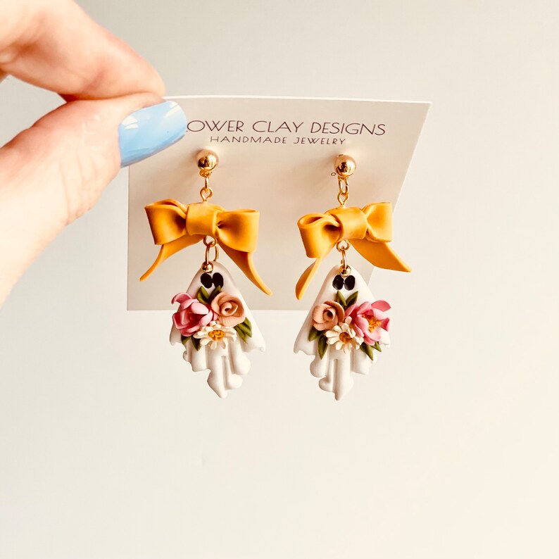 May include: A pair of handmade earrings featuring a white ghost design with a yellow bow and floral accents. The earrings are suspended from gold-colored posts. The card behind the earrings reads "Flower Clay Designs Handmade Jewelry."