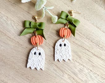 May include: A pair of Halloween-themed earrings. Each earring features a green bow, an orange pumpkin, and a white ghost with a floral pattern. The earrings have gold-colored posts and connecting hardware. The background is a light wood surface.