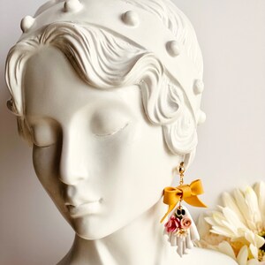 May include: A pair of gold-toned earrings featuring a ghost-shaped charm. Each charm has a mustard yellow bow and a cluster of pink and white flowers. The earrings are displayed on a white bust.