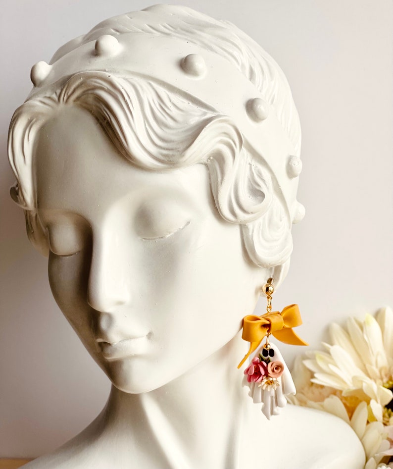 May include: A pair of gold-toned earrings featuring a ghost-shaped charm. Each charm has a mustard yellow bow and a cluster of pink and white flowers. The earrings are displayed on a white bust.