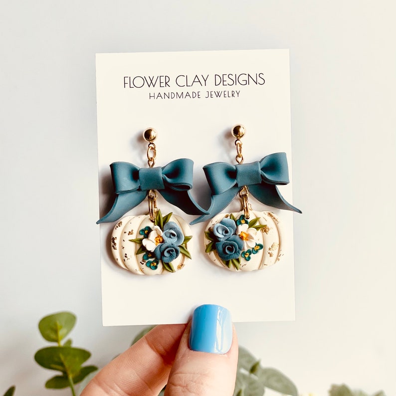 May include: Handmade earrings with teal bows and cream pumpkin charms decorated with blue and white flowers. The earrings hang from gold-coloured posts. The card behind the earrings reads "FLOWER CLAY DESIGNS HANDMADE JEWELRY."