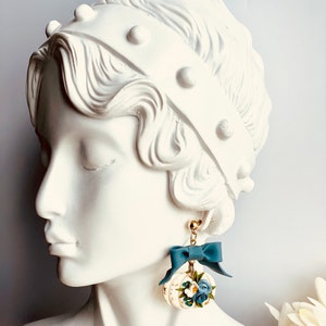 May include: Earrings with a white pumpkin design, gold detailing, blue flowers, and a matching bow. The earrings hang from gold-coloured posts. A delicate and elegant accessory, perfect for adding a touch of charm.