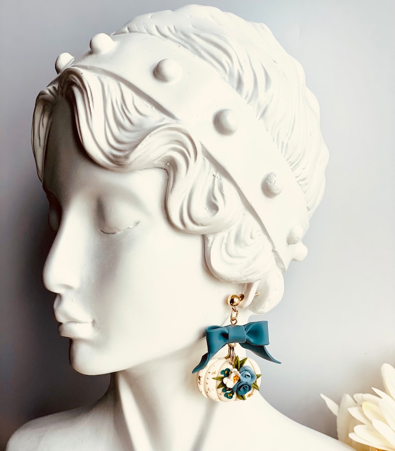 May include: Earrings with a white pumpkin design, gold detailing, blue flowers, and a matching bow. The earrings hang from gold-coloured posts. A delicate and elegant accessory, perfect for adding a touch of charm.