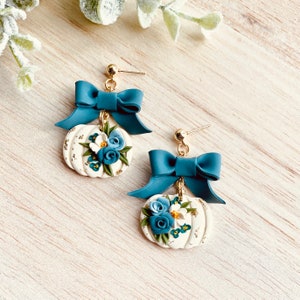 May include: A pair of handmade earrings. Each earring features a white pumpkin-shaped pendant with blue and white floral designs and gold accents. They have teal bows and gold-coloured posts.
