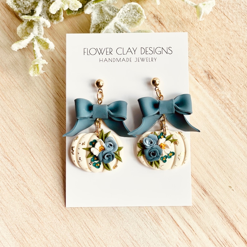 May include: Handmade earrings with a pumpkin design. The earrings have a cream-coloured pumpkin base with blue and white floral accents and gold-coloured leaf details. A blue bow tops each pumpkin, with gold-coloured hardware.