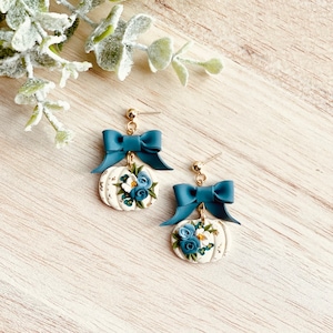 May include: Gold-coloured earrings with a teal bow above a white pumpkin charm. The pumpkin is decorated with blue and white flowers and gold accents. The earrings are set against a light wood grain background.
