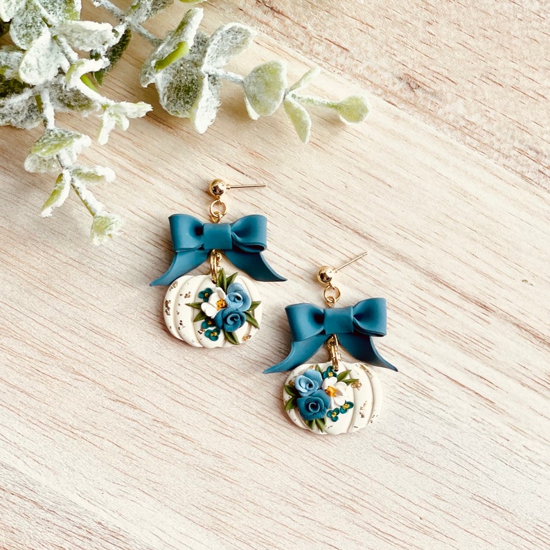 May include: Gold-coloured earrings with a teal bow above a white pumpkin charm. The pumpkin is decorated with blue and white flowers and gold accents. The earrings are set against a light wood grain background.