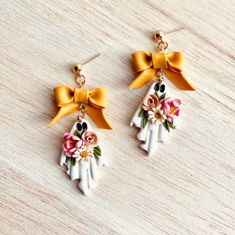 May include: A pair of handmade ghost earrings. Each white ghost-shaped earring features a cluster of pink, cream, and green flowers. A mustard yellow bow sits above each ghost, connected by a gold-colored post.